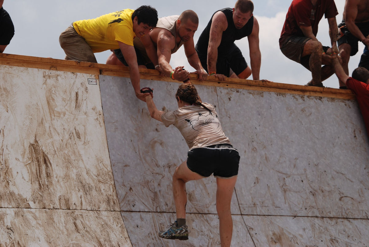 People helping a woman get up an obstacle course wall