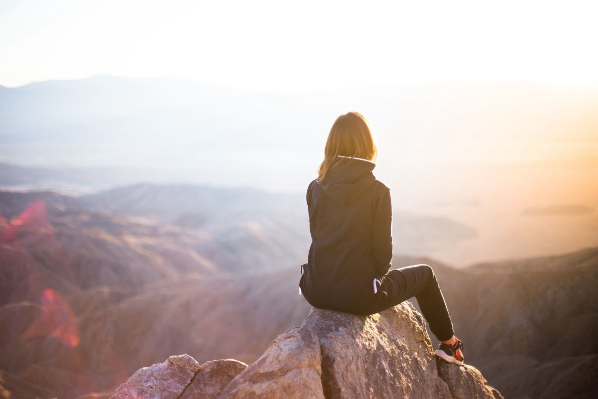 A woman sitting on top of a mountain pinnacle watching the sun rise in triump